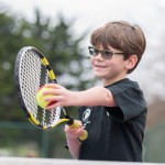 A young boy wearing glasses playing tennis with a black and yellow tennis racket.