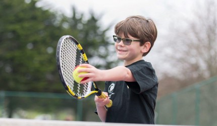 A young boy wearing glasses playing tennis with a black and yellow tennis racket.