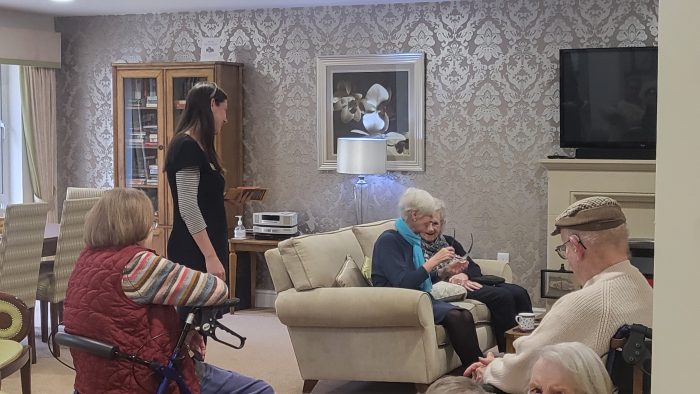 A woman wearing an iSightCornwall name badge stands in front of four older people who are seated on a sofa. They are listening to the iSightCornwall team member as holds a piece of technology and is talking to them.