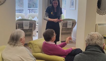 A woman wearing an iSightCornwall name badge stands in front of three older people who are seated on a sofa. They are listening to the iSightCornwall team member as holds a piece of technology and is talking to them.
