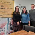Three people pose for the camera and smile. Behind them is a sign on the wall that reads Raison Opticians and a banner with the iSightCornwall logo and more information about the charity on.