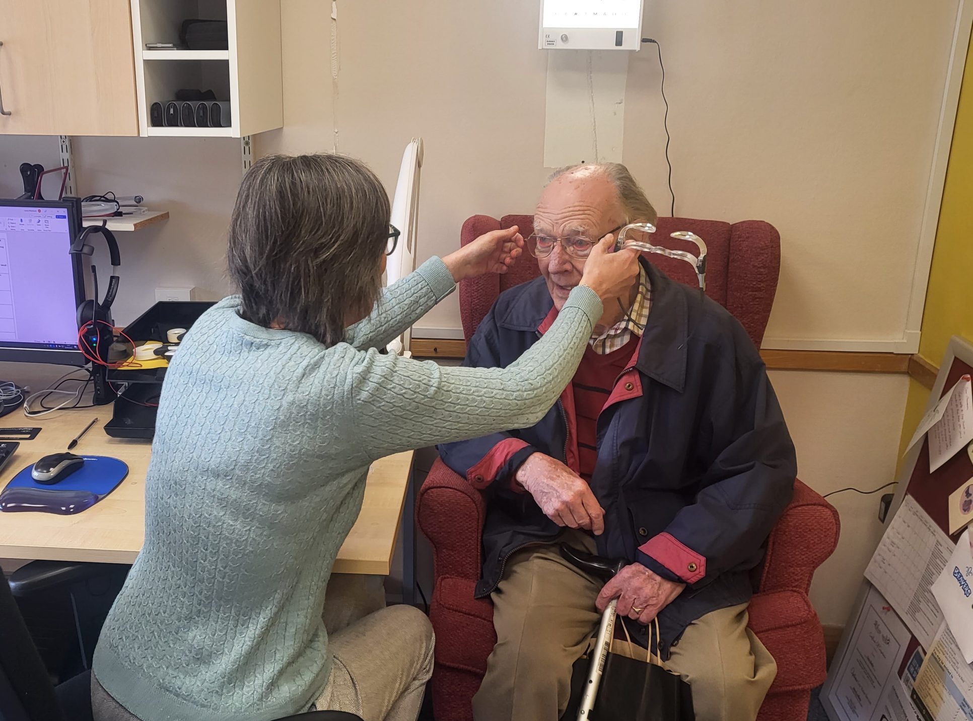 A man sits in a comfy chair looking at a woman. The woman is holding some MaxTV glasses in her hand and is adjusting the man's glasses on his face.