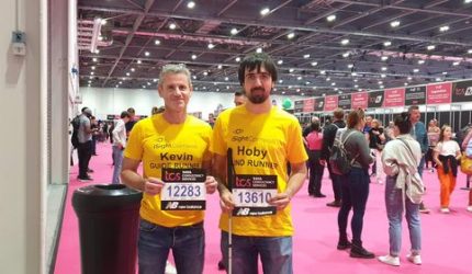 Hoby Allen (right) poses with guide runner Kev in the sign in hall of the London Marathon. They are both wearing their yellow iSightCornwall tshirts and are holding up their running number.