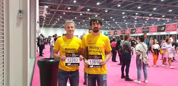 Hoby Allen (right) poses with guide runner Kev in the sign in hall of the London Marathon. They are both wearing their yellow iSightCornwall tshirts and are holding up their running number.