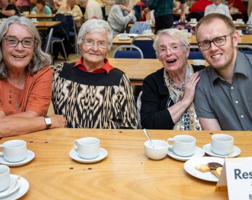 three women and a young man pose for the camera. They sit at the table and have cups of tea and coffee in front of them. They are all smiling.