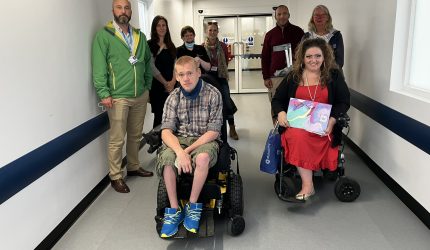 the members of the accessibility advisory group, making eight people in total, pose in the hallway of Treliske Hospital. Two are in wheelchairs, one is holding a symbol cane.