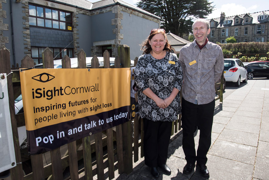Rod and Lynda from iSightCornwall stand in front of a sign advertising an iSightCornwall sight loss awareness event.