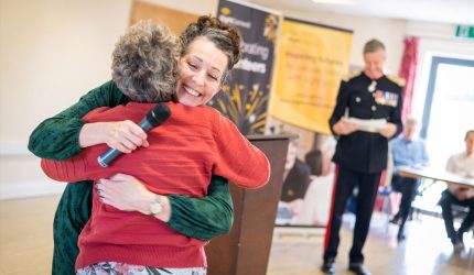 iSightCornwall Chief Executive, Carole hugs a volunteer as they are presented with an award at the recent AGM. Colonel Bolitho OBE stands in his uniform in the background.
