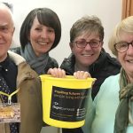A man wearing an iSightCornwall lanyard poses with three women holding an iSightCornwall bright yellow fundraising bucket. They all smile for the camera.