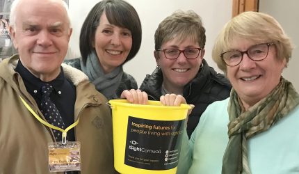 A man wearing an iSightCornwall lanyard poses with three women holding an iSightCornwall bright yellow fundraising bucket. They all smile for the camera.