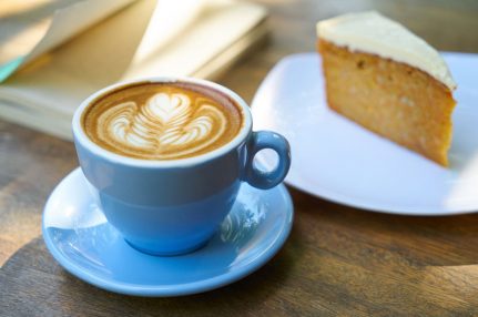 A latte coffee with a pretty coffee art pattern sits next to a plate with a slice of Victoria Sponge cake.