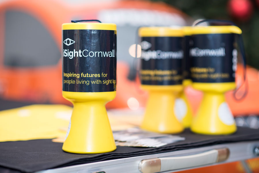 Four black and yellow, iSightCornwall branded collection tins on top of a silver fold-out table.