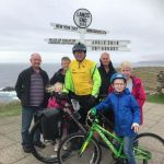 Craig Hancock wearing a yellow iSightCornwall vest on a bike, pictured with his family below the Land's End sign in Cornwall.