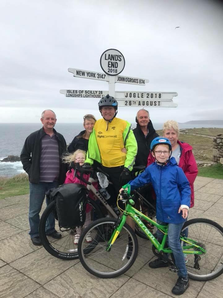 Craig Hancock wearing a yellow iSightCornwall vest on a bike, pictured with his family below the Land's End sign in Cornwall.