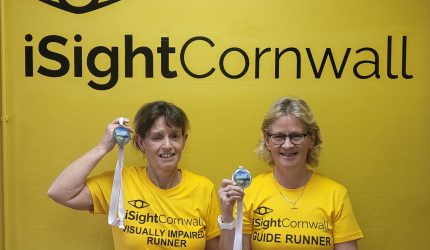 Image showing two women posing underneath a large iSightCornwall logo printed on the wall. They are both wearing bright yellow t-shirts which also have the iSightCornwall logo on, one says visually impaired runner and the other says guide runner. Both women are holding up medals next to their head and smiling.