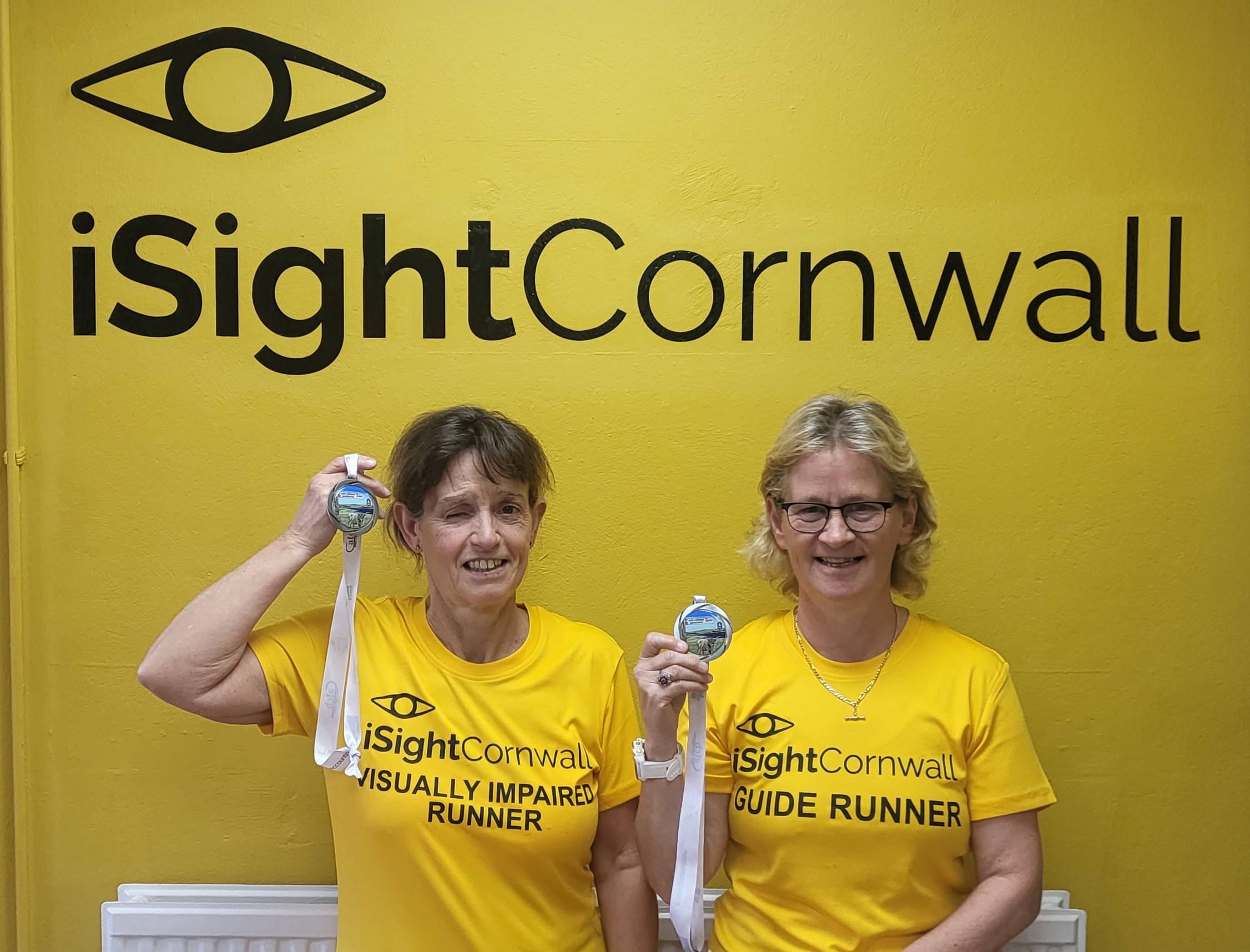 Image showing two women posing underneath a large iSightCornwall logo printed on the wall. They are both wearing bright yellow t-shirts which also have the iSightCornwall logo on, one says visually impaired runner and the other says guide runner. Both women are holding up medals next to their head and smiling.