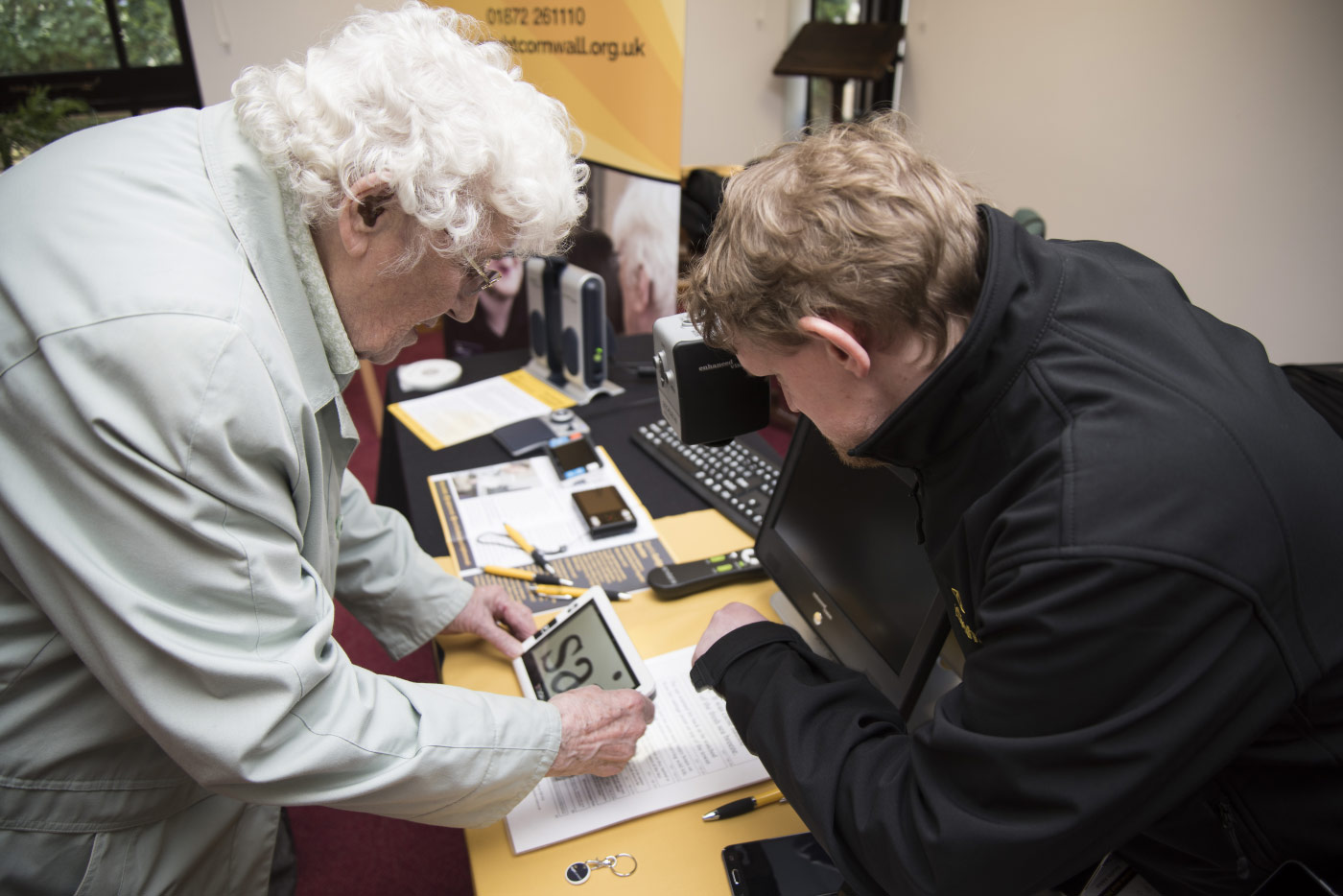 Dominic from iSightCornwall demonstrates a portable handheld magnifier to a lady in a white coat.