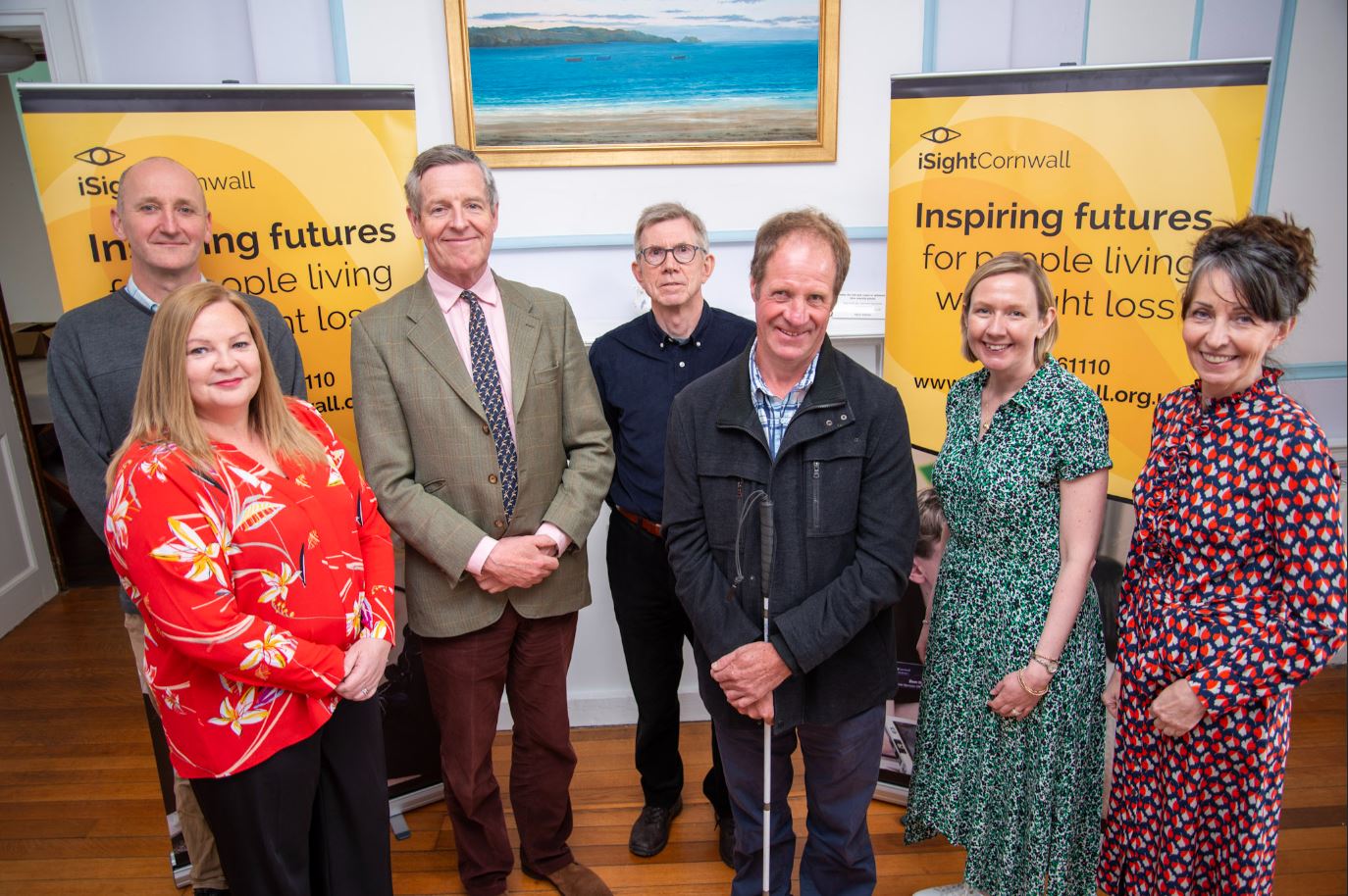 Seven people stand in front of bright yellow iSightCornwall banners, smiling at the camera.