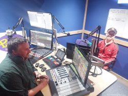 A young man sits in front of a mic inside a radio booth which is branded with Pirate FM's purple colours and logo. Opposite him, on the operative's side is another man who is looking at a screen and pushing a button,