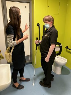 iSightCornwall team members, Dom and Jodi, inspect the accessible toilet at the hospital. The walls are a bright yellow and the handles black. Jodi is gesturing to one of the handles whilst Dom stands listening whilst holding his white symbol cane.