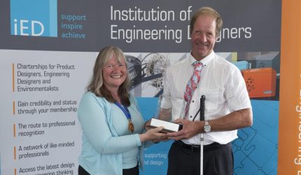A man poses holding a clear glass cube engraved on a small podium. He also has a white cane in his hand. He smiles as he holds the cube shaped award alongside another woman. They stand in front of a banner which reads institution of engineering designers.