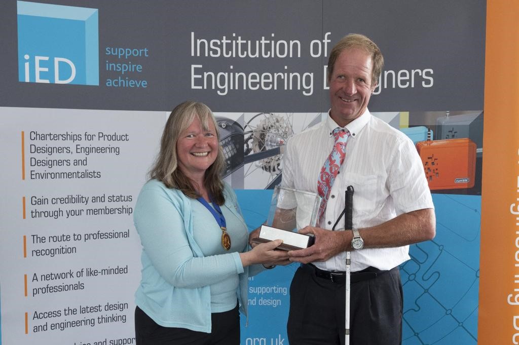 A man poses holding a clear glass cube engraved on a small podium. He also has a white cane in his hand. He smiles as he holds the cube shaped award alongside another woman. They stand in front of a banner which reads institution of engineering designers.