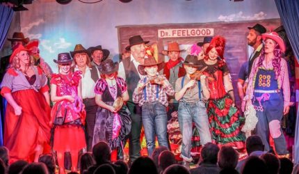 A group of about 20 people stand on a stage dressed in fantastical clothes and cowboy hats as an audience watches on