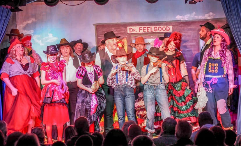 A group of about 20 people stand on a stage dressed in fantastical clothes and cowboy hats as an audience watches on