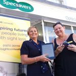 iSightCornwall's, Lynda, stood outside Specsavers Opticians with a member of the team, both ladies are smiling holding glasses and a tablet.
