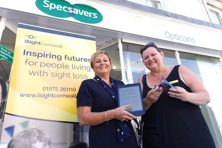 iSightCornwall's, Lynda, stood outside Specsavers Opticians with a member of the team, both ladies are smiling holding glasses and a tablet.
