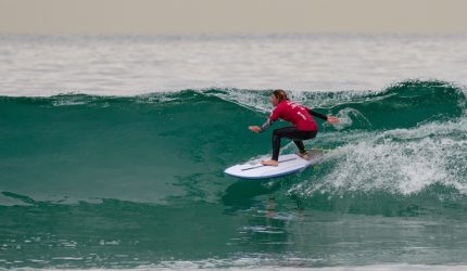 Melissa Reid surfing a wave on a white surf board wearing a red top.