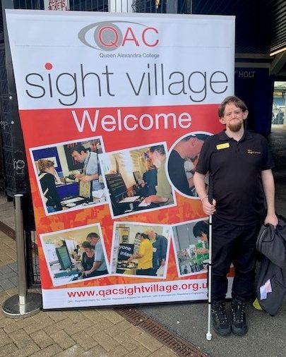 A young man wearing all black with an iSightCornwall staff badge that reads 'Dominic Hall' stands holding a white cane. Behind him is a large banner for the Sight Centre event.
