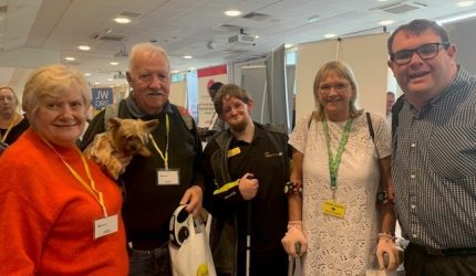 iSightCornwall's Dom Hall poses with some iSightCornwall volunteers and clients. All together 5 people are posing and smiling for the camera, four are wearing yellow lanyards whilst Dom in the middle is wearing his iSightCornwall staff badge and is holding his white cane.