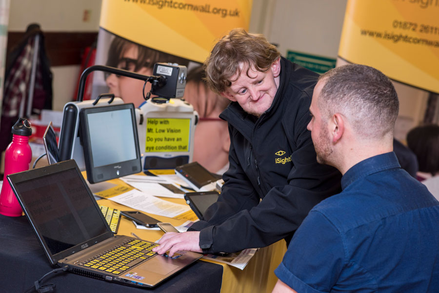iSightCornwall Assistive Technology Officer, Dom Hall demonstrates a specially adapted laptop to a client. Various assistive technology devices can be seen in the background.