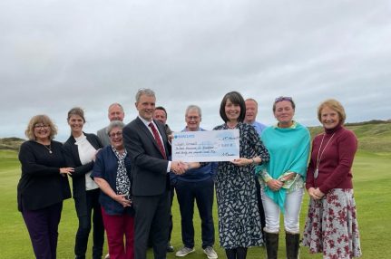 Members of the golf club stand on the green smiling for the camera. In front of them stands a man in a suit holding a cheque. On the other side is a smiling woman accepting the cheque.
