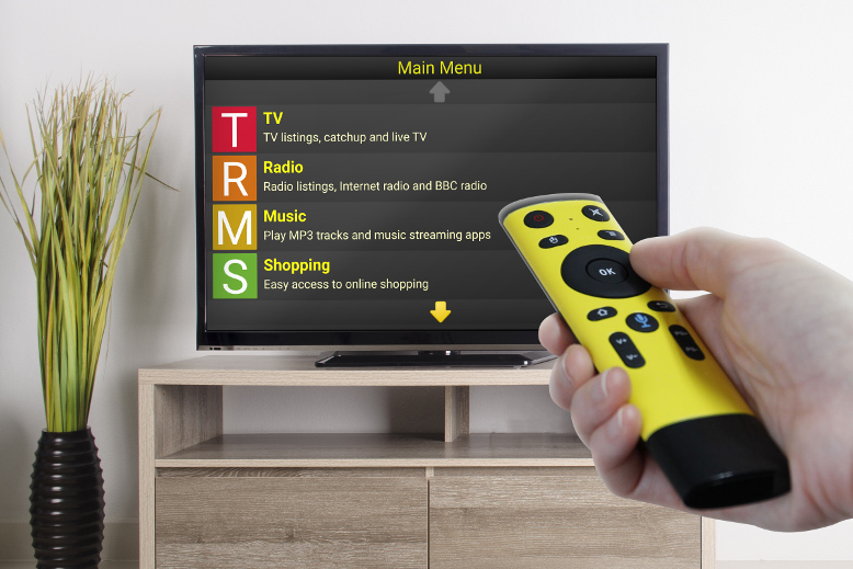 The Synapptic TV box is displayed on a flat screen television. A bright yellow remote control is pointing directly at the television.