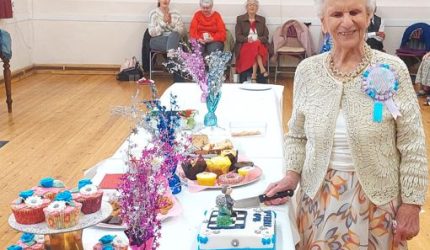 A woman stands next to a table full of cakes, decorations and other food. In her hand is a knife which is resting against a birthday cake which features to figures made up of icing. The woman is wearing a blue rosette which says it 's my birthday.