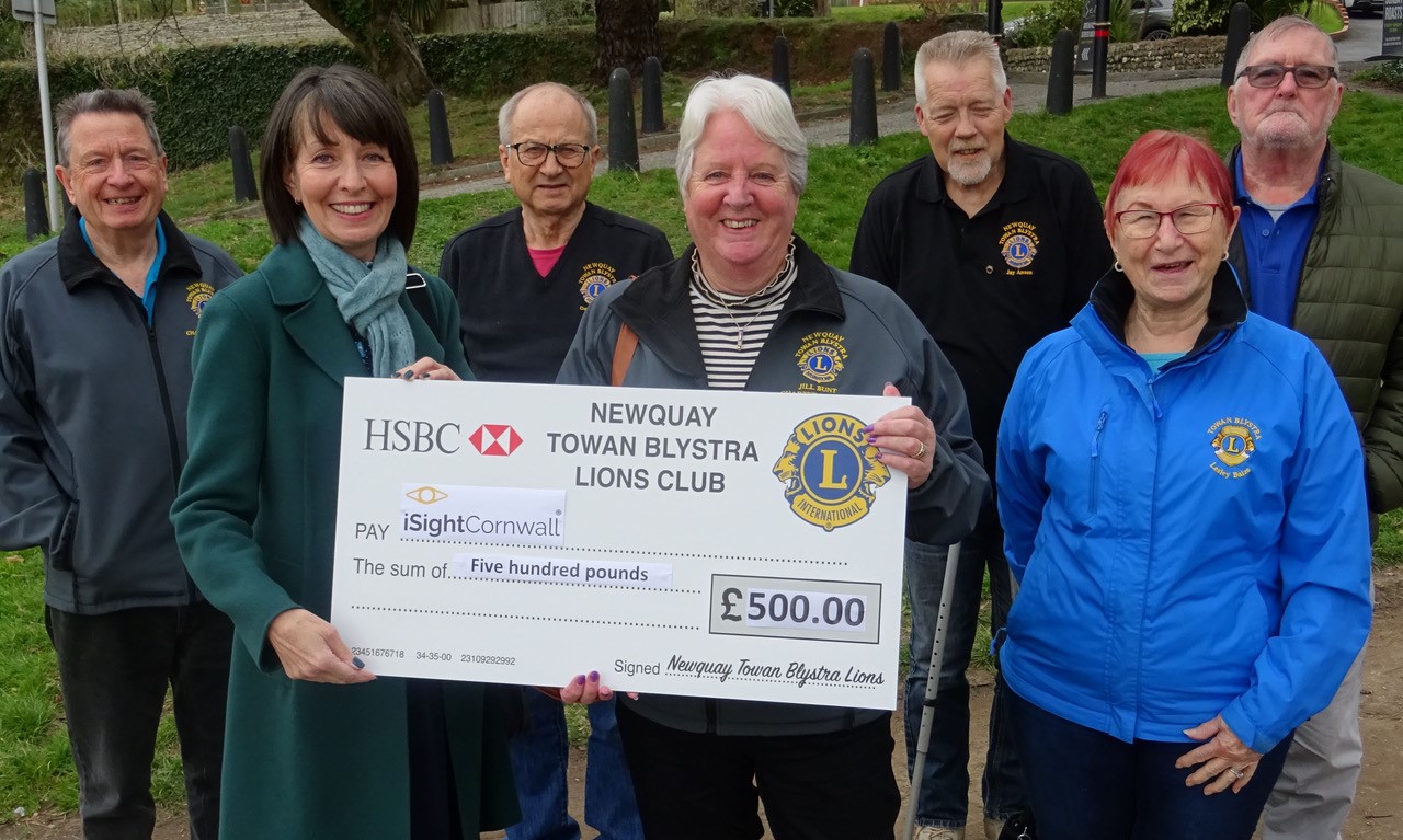 A group of people wearing matching jackets with the Lions Club logo on the left hand side stand smiling as two woman stand in the front holding a giant cheque.
