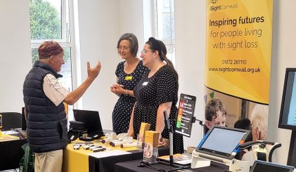 Two women stand behind an iSightCornwall branded table. They listen intently to a man in front of them who is gesturing to the magnifiers on the table.