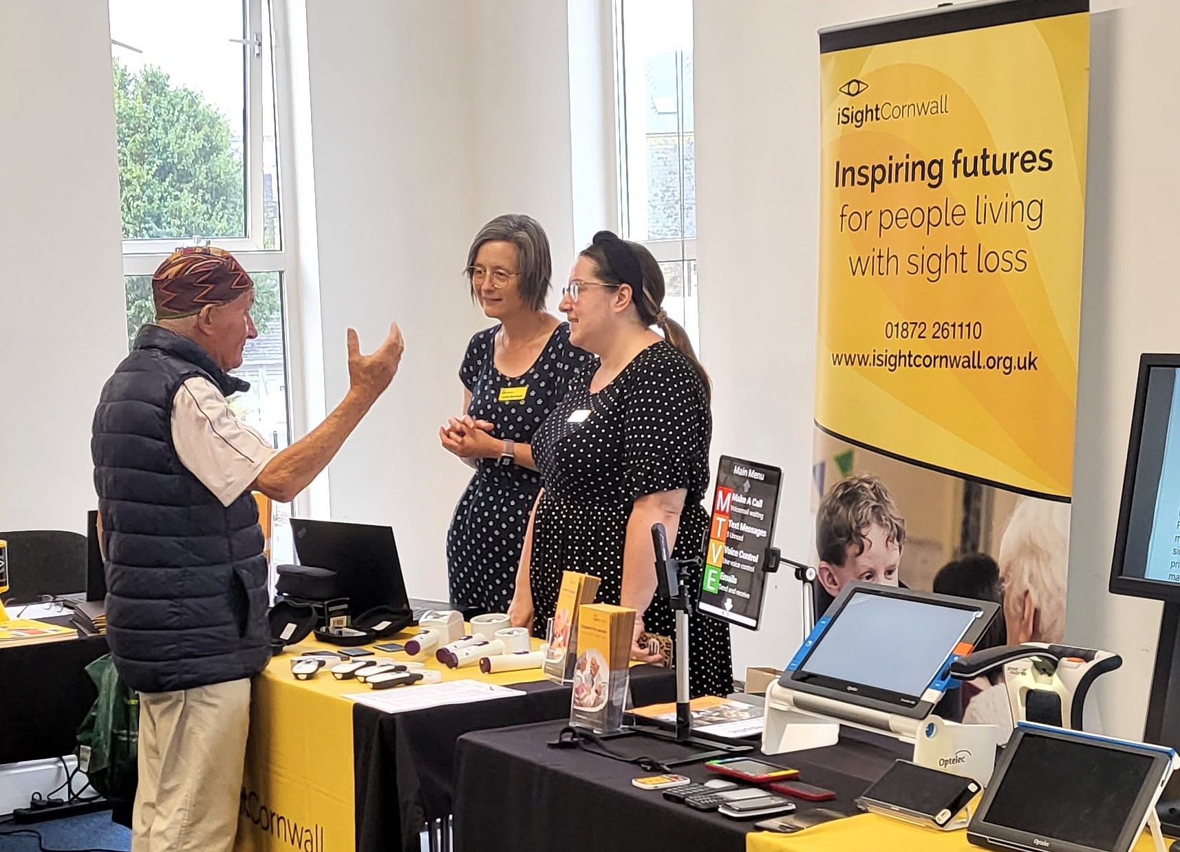 Two women stand behind an iSightCornwall branded table. They listen intently to a man in front of them who is gesturing to the magnifiers on the table.
