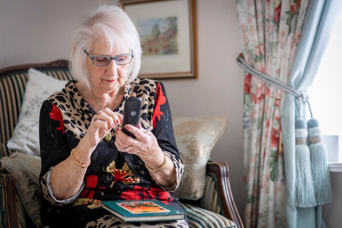 The image shows a woman sitting in an armchair in a cosy home as she looks down at the landline phone handset in her hand and pushes a button.