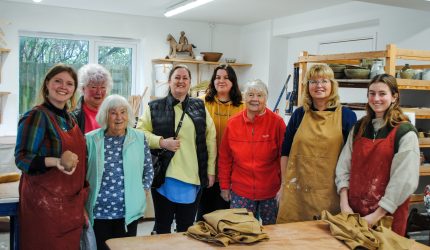 A group of women all stand for a photo in a pottery. Some are wearing aprons. They are surrounded by shelves with sculptures, cups and other clay creations on them.