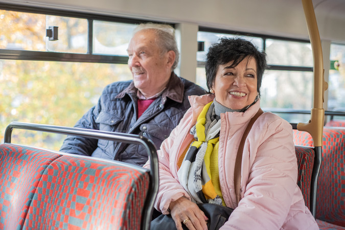 A man and a woman sit next to each other on a bus. The woman is smiling and looking off camera.