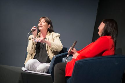 iSightCornwall's CEO, Terri Rosnau-Ward, in a cream jacket talking to a crowd whilst holding a microphone. A lady in a red jumper holding a microphone is sat next to her.