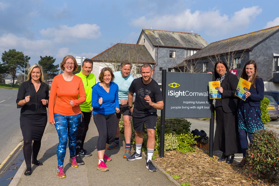 Six people dressed in running gear pose outdoors next to the iSightCornwall sign on a sunny day.