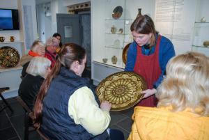 A woman holds a large decorative plate out to another woman who is sitting down. She is reaching out with her hands to feel the design on the plate.