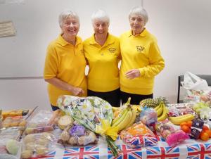 Three women wearing bright yellow jumpers with the Saltash BAPs logo on are smiling and have their arms around each other as they pose for a photo. In front of them is a table full of food and harvest produce.