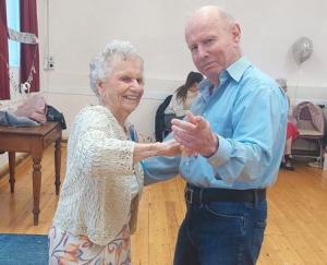 A woman wearing a birthday rosette holds hands with a man as they dance, smiling at the camera.