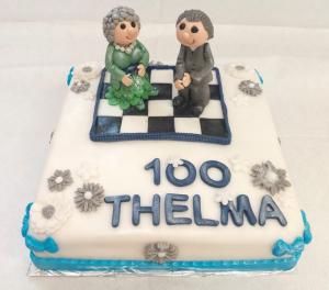 A birthday cake with the words '100 Thelma' and two figures dancing on a checkered dance floor. One of the figures looks like Thelma, the birthday girl.