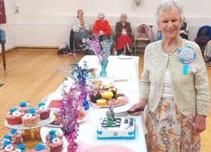 A woman wearing a birthday rosette stands next to a table of food and decorations. She is holding a knife which is resting against a birthday cake.